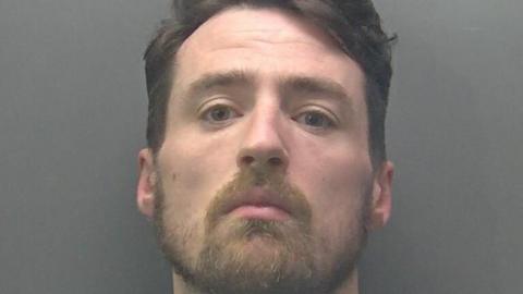 A custody photo of Mark Moss, who is positioned against a plain, grey background. He has short, dark hair that appears neatly styled, with the top slightly voluminous and the sides trimmed closer. His ears are visible on both sides.