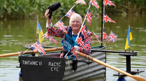 An elderly man in a home-made boat made from corrugated metal. He is waving his dark blue cap while wearing a Union Jack jacket, with Union Jack and Ukrainian flags on the boat.