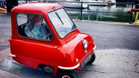 A red Peel P50 is parked up close to the harbour in Peel. A man in a white hat and blue shirt is in the small car and has one hand on the driving wheel.