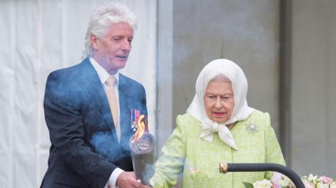 Bruno Peek in dark suit and thick white hair hands a flaming torch to the late Queen Elizabeth II, who is wearing a white headscarf and lime green coat. A large bouquet of pink, white and lilac flowers are in front of her. 