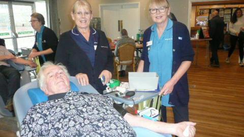 Dave Charles - a man with white hair, wearing a patterned t-shirt and navy trousers, sits in a blue chair. He is holding a blood bag. Two women in healthcare uniform are standing behind the chair. They are indoors in a room with wooden floors and neutral-coloured walls.