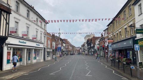 A shot of a traffic free main road in Farnham showing shops and bunting across the road. There are a handful of pedestrians on the pavements on each side of the road. 