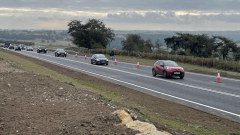 Vehicles travelling on a newly laid road with red and white cones lining the road on the far right of the picture. To the left of the picture is soil and a few light brown stones with short green grass.