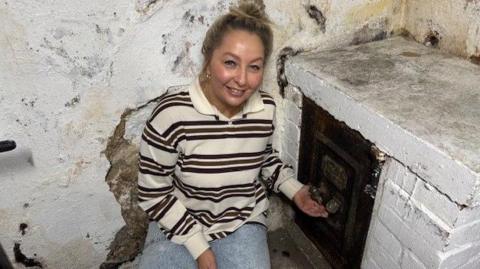 Lyndsey Guthrie is in the pub cellar which is white bricked. She is crouching down beside the safe which is set into the wall. She is a white jumper with dark stripes. She is smiling and her blond hair is in a bun. The safe is dark brown and rusted.