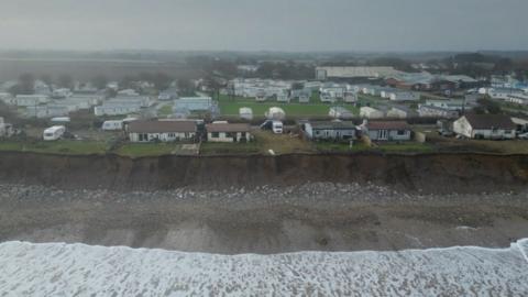 Holiday chalets and caravans are close to the edge of a cliff-top. Below them is a sandy beach with white stones and the tide coming in, which is foamy and white.