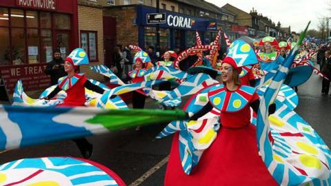 People are dressed in colourful red costumes and elaborate turquoise head gear. They are dancing down a busy street as part of a parade at a fair