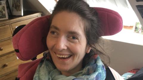 A brunette woman wearing a blue scarf smiles at the camera. Behind her is a read headrest, a windowsill and a chest of wooden drawers.  