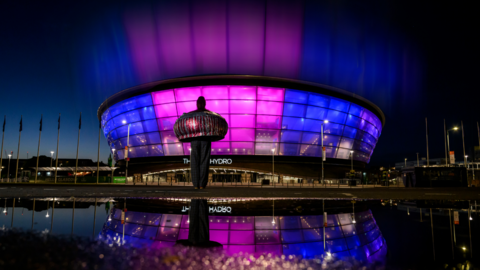 The OVO Hydro arena in Glasgow is lit in purple and blue at night, reflected in a puddle in the foreground, with a large sculpture standing outside the building.