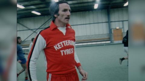 Derek Dougan wears a red and white Kettering Town football top with Kettering Tyres printed on it