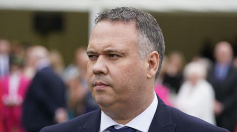 A man with short grey and black hair looks off to the side of the camera. He is wearing a dark-coloured suit jacket, a white shirt, and a tie. The background is out of focus, but features a number of people standing. 