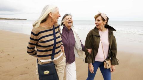 Three women smile and laugh together while walking on a beach under a grey sky.
