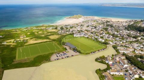 An aerial view of a two football fields and housing in St Ives. The pitch on the left is bigger and is horizontal and the one on the right is vertical.  The sea is in the background. 