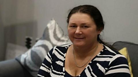 A woman with brown hair sits on a grey sofa. She wears a black and white striped top. Her hair is tied back into a ponytail.