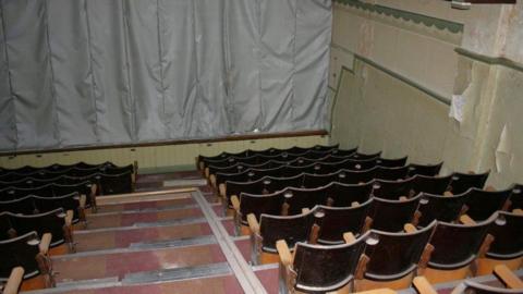 An interior shot of the original cinema inside. It shows a dark room with rows of dusty wooden chairs - four either side of the central steps - which leads down to the screen which is covered by a grey tarpaulin. 
