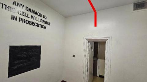 The interior of a holding cell with white-painted walls and a white ceiling. A doorway on the right-hand side leads into an adjoining corridor.
A thick red line has been digitally overlaid onto the image, running across the ceiling and down the wall above the doorway. Large black lettering painted onto the wall reads: ANY DAMAGE TO THIS CELL WILL RESULT IN PROSECUTION.