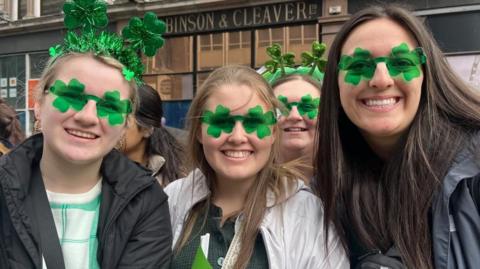 Three women smile into the camera wearing green shamrock sunglasses.
