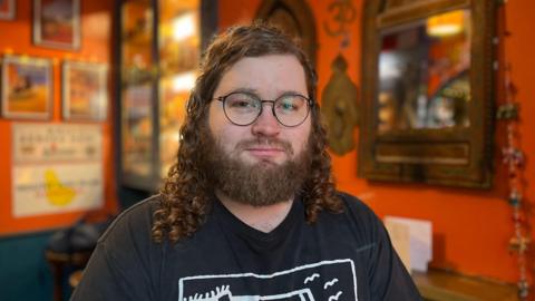 George Taylor, the pub's owner, who has long curly hair, a beard and glasses looking pensively at the camera. He is wearing a black t-shirt with a white logo on the front and standing in one of the rooms of the pub which is painted orange with pictures and a mirror hanging up 