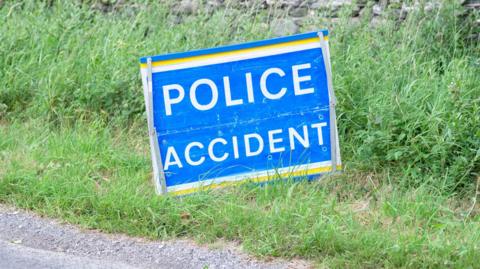 A police accident sign with white lettering on a blue background on a grass verge near a road
