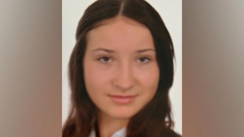 A teenage girl with long brown hair looks at the camera. She is wearing a white shirt, a navy and white striped tie and a navy blazer.