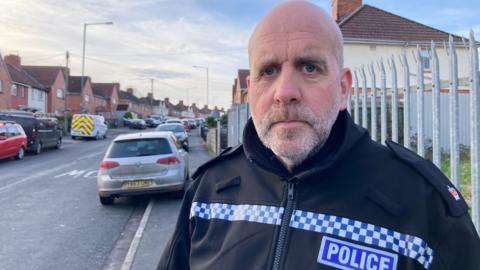 Supt Mark Runacres. He is bald but has light grey stubble on his chin and upper lip. He is pictured looking at the camera with a slight frown. He is wearing a black police jacket with a blue police logo on the chest. 