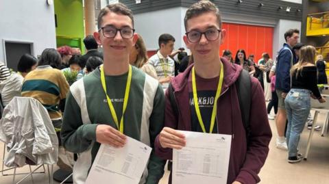 Identical twins, both with glasses, one wearing a green jumper and one in a red jacket. Both are holding up their A-level results. 
