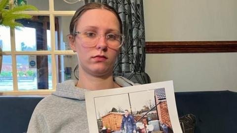 Kayce (pronounced Casey) Burton is sitting on a blue sofa, holding a picture of her dad. He is on a building site, where he had been fitting windows. He is wearing blue jeans and a blue jacket, and there is a shed and tools and the back of a house in the photograph. Kayce is wearing a grey hoodie, and glasses, and has her hair tied back in a ponytail. There is a window behind her, with blue curtains and a plant pot with a floral display.