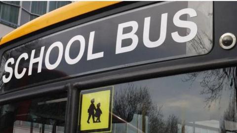 Image shows the front windscreen of a school bus with the words school bus written at the top of the windscreen and in the middle of the windscreen at the top is a shadow picture of two children with a yellow background. 