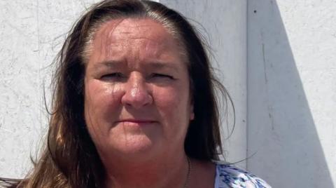 Marion Atkinson, with long brown hair and a white, pink, green and black speckled top, stands in front of a white wall and looks into the camera