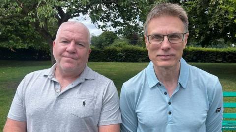Simon Byrne and Adam Rainford are standing side by side in a park. Simon is wearing a grey polo short while Adam is wearing a blue polo shirt.