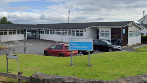 A one storey white building with a black roof and windows all around the exterior. In front of it is a car park which has a black and red car in it. In front of that is a grass bank with a blue and silver sign saying 'Fairhill Medical Practice'.