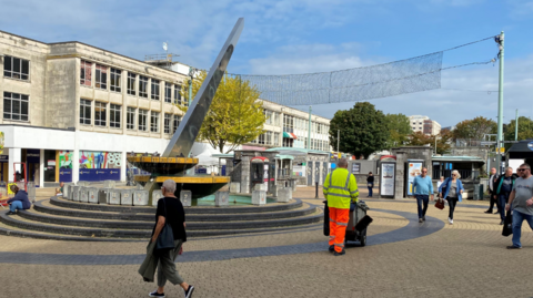 A large sundial in a city shopping centre with people including a council operative in hi-vis walking around it. It is a sunny day with blue skies and light cloud cover.