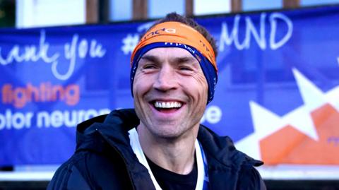 Kevin Sinfield - a man wears an orange headband with MND written on it in white and blue fonts while smiling. He is stood in front of a large blue MND banner.