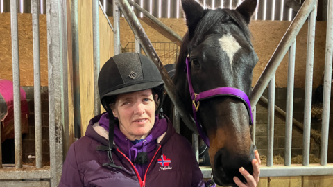 Lesley Sayers standing in a stable with her horse Seb. She wears a riding cap and purple coat and strokes Seb's nose