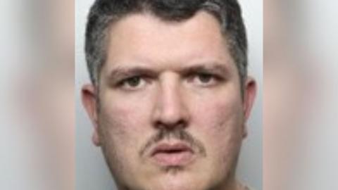 A mugshot of a frowning man with a small mouth, moustache and soul patch. 