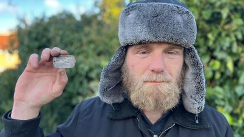 A man wearing a russian style bearskin hat with fluffy ear coverings holds up a metallic military dog tag. He has a mid-length blonde beard and wears a blue jacket. He is standing in front of high green bushes, which are out of focus.