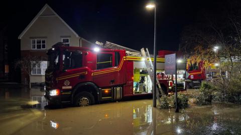A flooded area in a neighbourhood with a red fire truck seen parked in the water next to a house. A street lamp is on with a "for sale" sign near a front garden which is flooded.