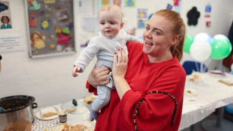 Kelly, wearing a red dress, smiles as she holds her baby