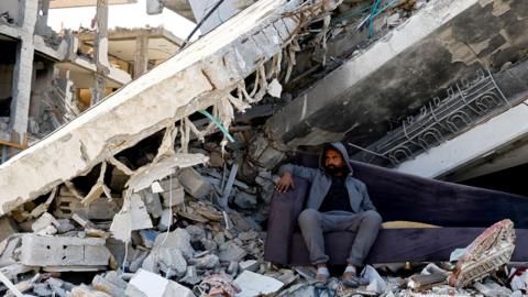 A Palestinian man sits on a purple sofa amidst the rubble of buildings destroyed during the Israeli offensive, amid the ceasefire between Israel and Hamas, in Rafah in the southern Gaza Strip