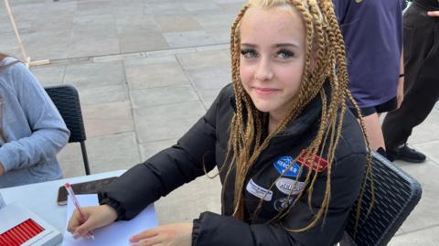 Neveah sitting at table in Broad Street in March. She is wearing a black jacket, has multiple long blonde plaits and is looking up and starting to smile. In her right hand is a pen, poised above paper. 