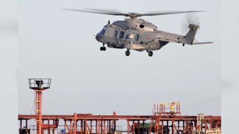 A grey helicopter seen above an orange and blue oil tanker