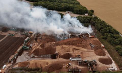 An aerial photos shows smoke drifting across a square site, with piles of bark and machinery located across the area. Trees and fields border the site. 