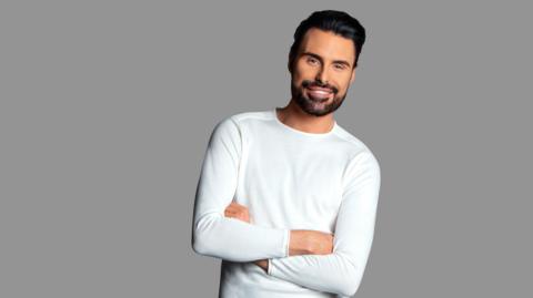 Rylan Clark-Neal stands with his arms crossed - he has short black hair and a beard