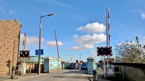 Sulcoates Bridge before its closure to traffic in 2020. It has light blue metal structure and barriers and warning lights at the end of the bridge. Two cars are driving across.