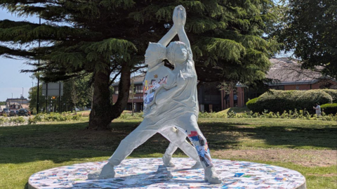 The grey sculpture, which is two people intertwined with their hands joined above their heads. Colourful patterns can be seen in sections through the sculpture. It is on a patch of grass with a road behind it.