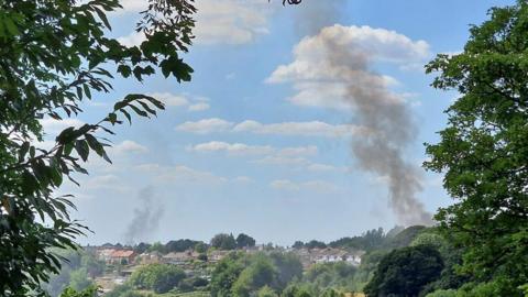 A view through tree branches of a sunny day and roofs of homes - a thick plume of smoke is on the right and slightly smaller one on the left 