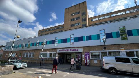 People walking into Yeovil District Hospital. The building has brown bricks and purple lettering saying 'Yeovil District Hospital'. There are cars outside and a zebra crossing.