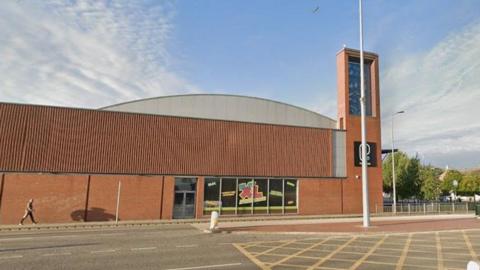 A Google street view of a brown building which is long and has a few windows on its side and a side door. A sign says 'Play XL' and another sign further along on a higher part of the building reads 'PureGym'. It is situated by a main road.