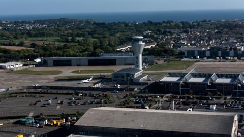 An aerial view of Jersey Airport which shows a large car park to the left with the glass airport building to the right with a triangular glass roof. Behind the airport building is the spherical shaped silver control tower. In the background are houses and trees and behind them is the sea.