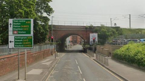 Dysart Road, looking towards traffic lights and a red-brick railway bridge. The road surface looks damaged and in the foreground a green sign gives directions to the town centre and other locations, including Boston, Nottingham and the train station.