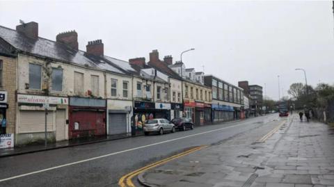 A wet street from the rain on a dark, drizzly say. On the left hand side of the road sits a row of empty shops with shutters boarded up. On the right hand side of the road, two people walk towards a bus stop.